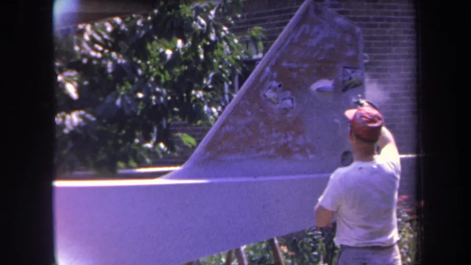 The man continues painting the aircraft tail fin in an outdoor Yucaipa setting, with trees and brickwork behind him.