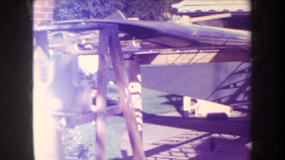 A wider view shows aircraft parts on wooden stands in the outdoor work area, with strong 8mm exposure flare.