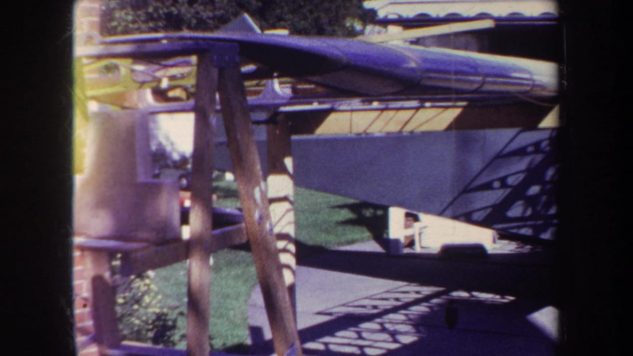 1969 Yucaipa home-movie frame showing a green biplane close to a garage, with fabric wing surfaces, wooden struts, lawn, and hard driveway shadows.