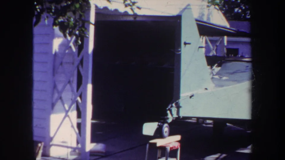 1969 Yucaipa home-movie frame showing a green biplane resting on a trailer outside an open garage, with a small stool, driveway, and lattice shadows visible.