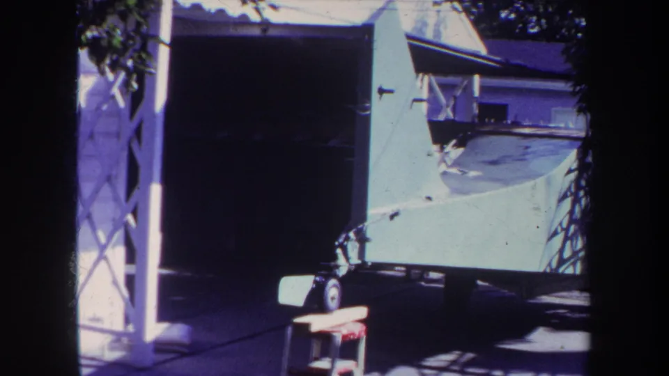1969 Yucaipa frame showing the biplane on a trailer outside a residential garage, with the wing surface, trailer wheel, stool, siding, and strong sun shadows.