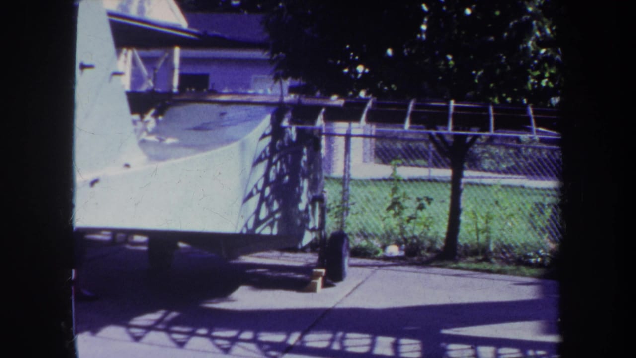 1969 Volmer, Yucaipa home-movie frame showing a chain-link fenced yard, concrete patio, green lawn, tree shade, and a large white structure at left.