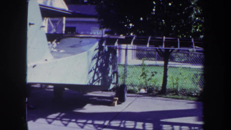 1969 Volmer, Yucaipa home-movie frame showing a chain-link fenced yard, concrete patio, green lawn, tree shade, and a large white structure at left.