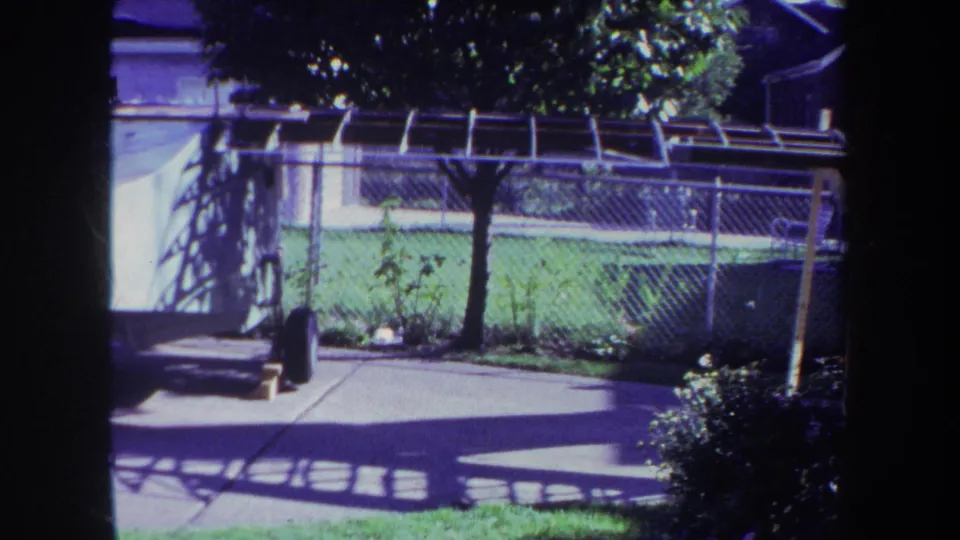 1969 Volmer, Yucaipa home-movie view of a residential yard, showing patio shadows, lawn, shrubs, chain-link fencing, and part of a white structure.