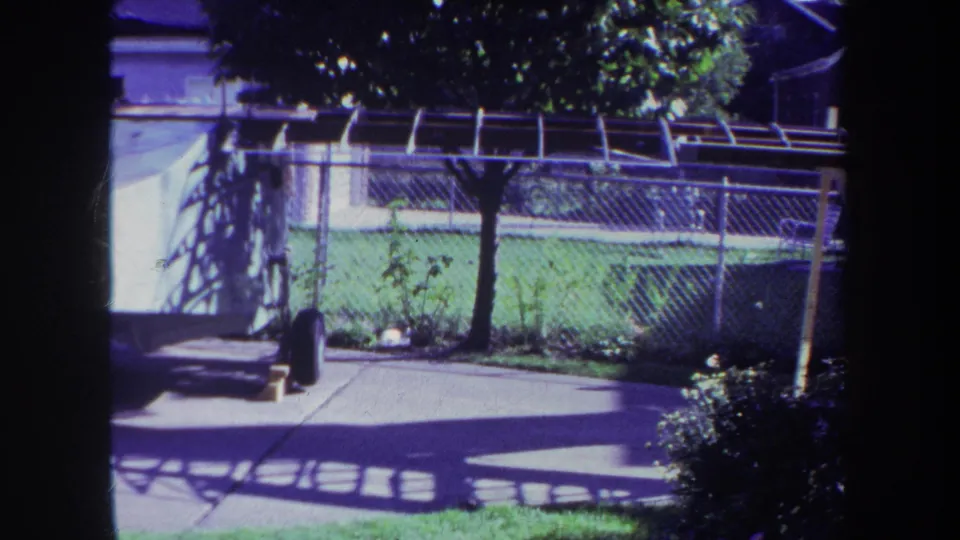 1969 Volmer, Yucaipa frame showing a residential backyard with chain-link fence, concrete patio, lawn, tree shade, and a large white structure on the left.
