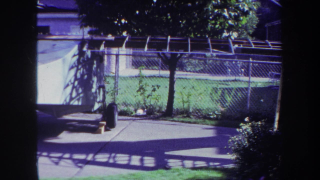 1969 Yucaipa home-movie frame showing a fenced backyard, covered pool area, lawn, patio shadows, and a white watercraft on a trailer.