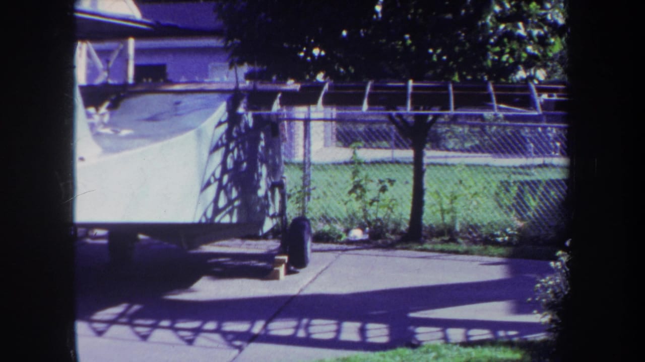 Fenced backyard with covered pool area, white watercraft on trailer, grass, trees, and house exterior.