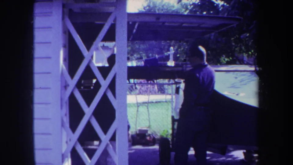 1969 Yucaipa home-movie frame showing a man from behind beside a boat, framed by porch lattice and backyard fencing.