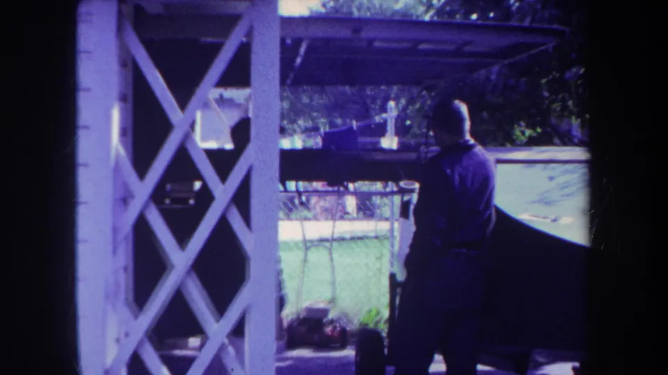 1969 Yucaipa backyard frame with a man near a boat, chain-link fence, lawn, and white lattice porch supports.
