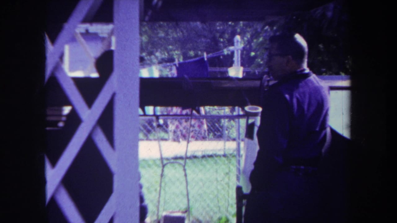 In 1969 Yucaipa home-movie footage, a man with glasses stands on a shaded porch, partly framed by white lattice and backyard fencing.