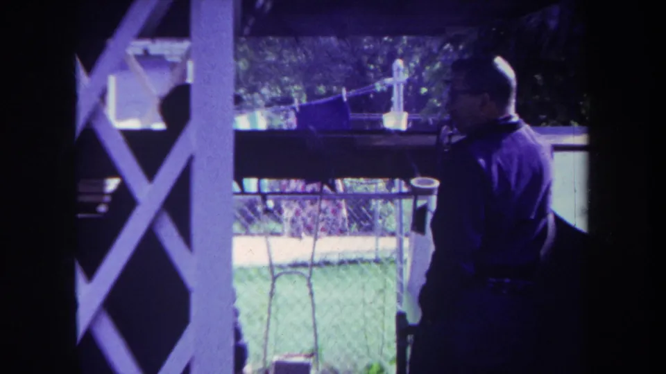 In 1969 Yucaipa home-movie footage, a man with glasses stands on a shaded porch, partly framed by white lattice and backyard fencing.