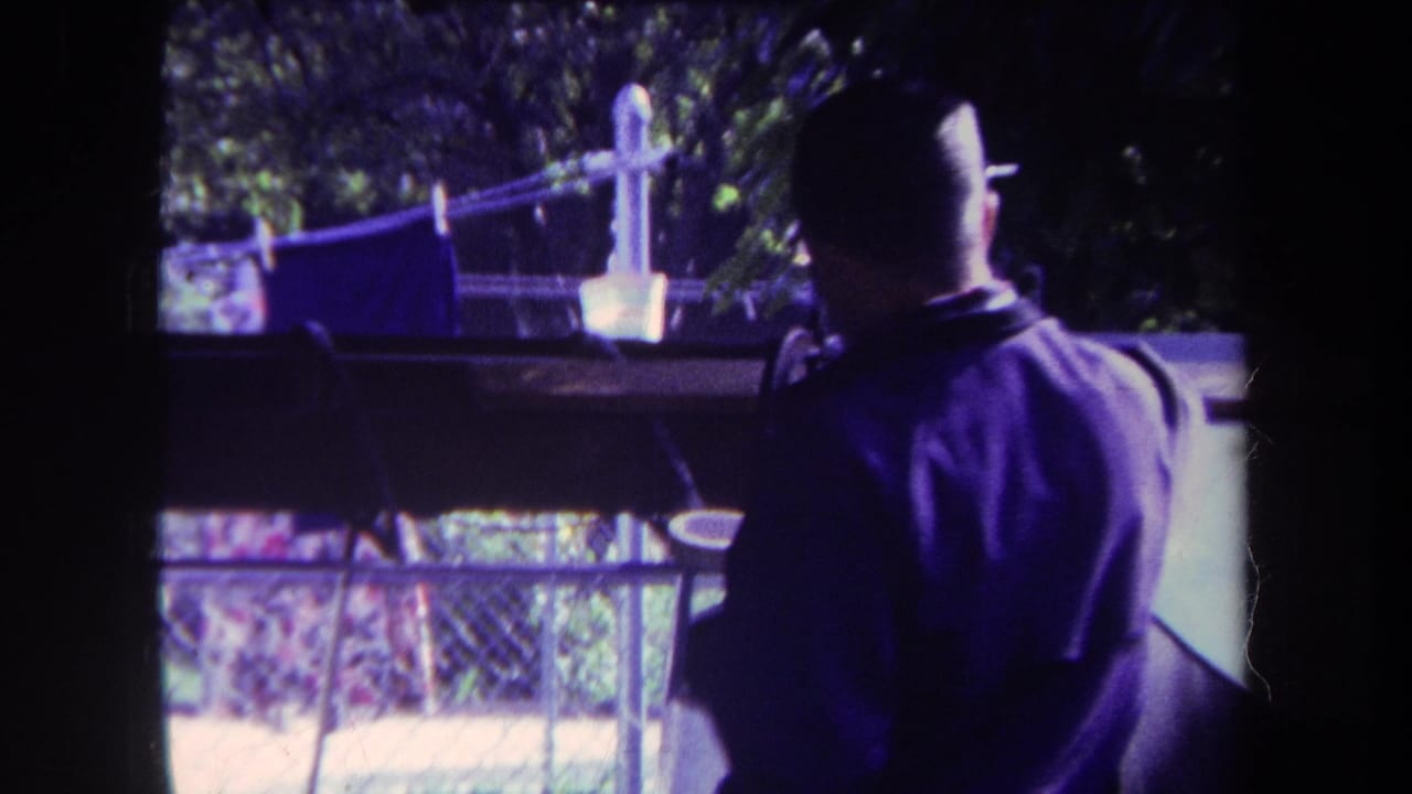 Man seen from behind on a porch, facing a backyard fence and laundry line in bright sun.