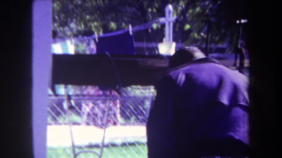 The man bends forward in the shaded porch area, with purple-tinted 8mm color, backyard fencing, and a clothesline behind him.