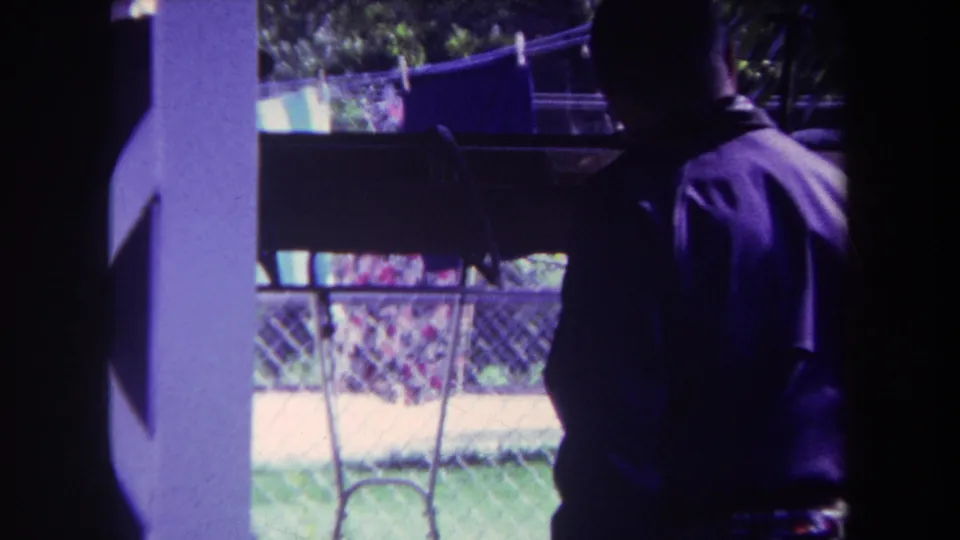 A side view shows the man on the shaded porch in 1969 Yucaipa footage, with a white post, clothesline, and chain-link fence behind him.