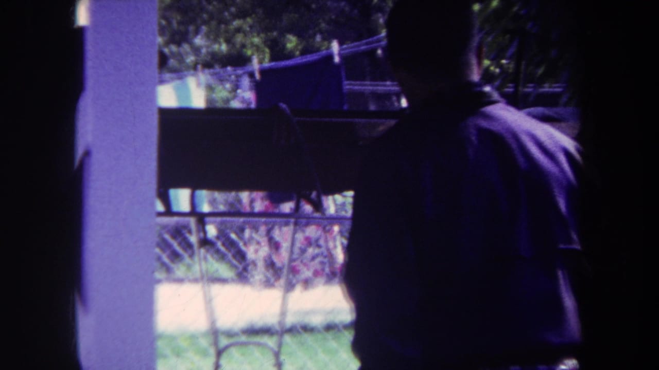 In 1969 Yucaipa home-movie footage, a man in a purple shirt stands indoors facing a backyard clothesline beyond a window or doorway.