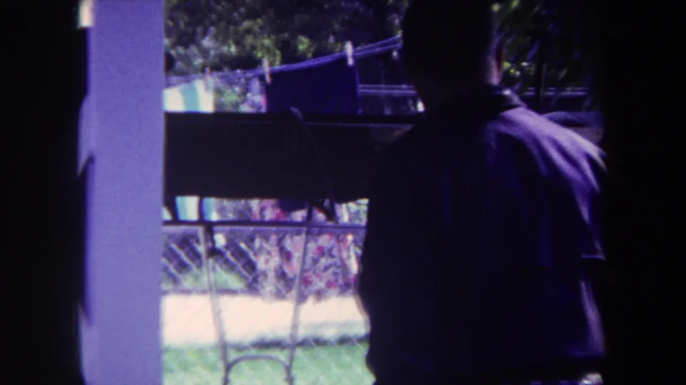 In 1969 Yucaipa home-movie footage, a man in a purple shirt stands indoors facing a backyard clothesline beyond a window or doorway.