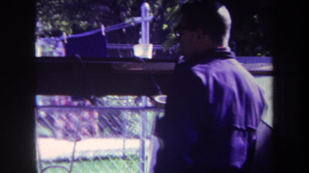 Man in a purple shirt stands near a dark window frame with laundry and yard visible outside.