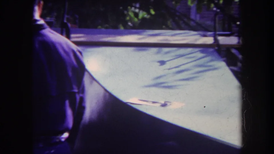 A partially visible man stands at the left edge of a shaded interior, facing a bright patio area and backyard fence in the home-movie scene.