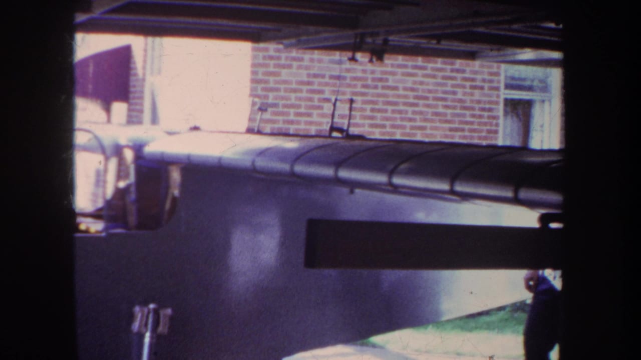 Large wooden model airplane under a covered structure with a red brick building behind it.