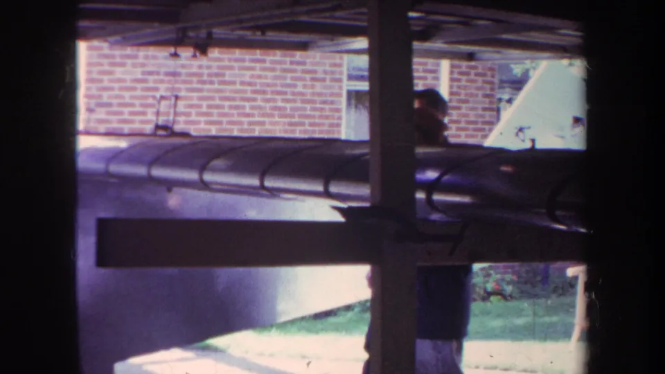 1969 Volmer Yucaipa frame showing a person beside the model airplane's forward section, with brick wall, beams, grass, and walkway visible.
