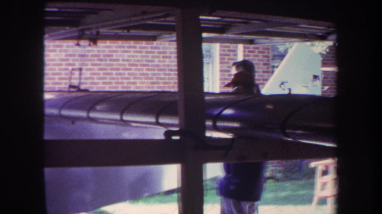 1969 Yucaipa home-movie frame showing a person partly obscured by a window crossbar, a dark cylindrical object, brick wall, and lawn.