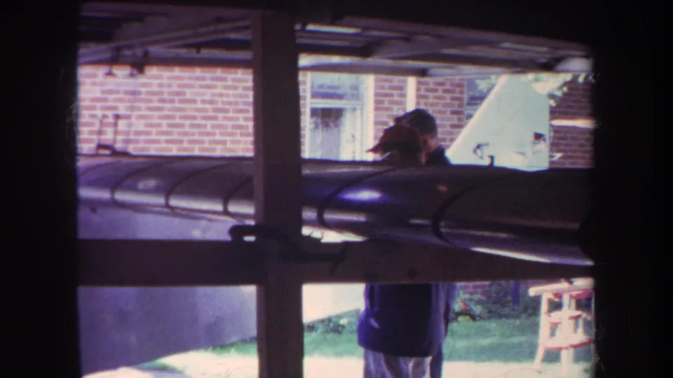 1969 Yucaipa frame with a cap-wearing person, brick house wall, lawn, and dark cylindrical object seen through window framing.