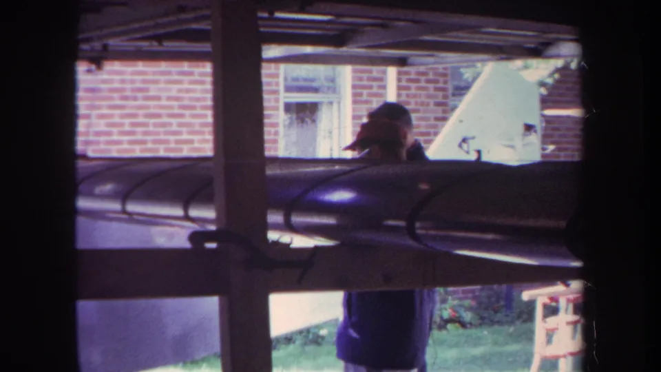 1969 Yucaipa home-movie still showing a partially visible person in a cap behind a dark object, with lawn and brick exterior beyond.