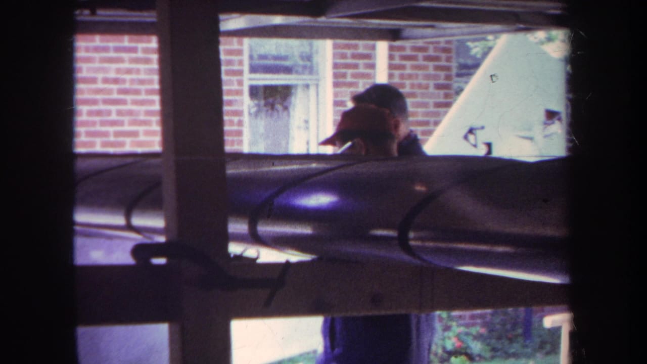1969 Yucaipa home-movie frame showing a man in a cap outside a brick house, partly blocked by window mullions and an aircraft wing or fuselage.