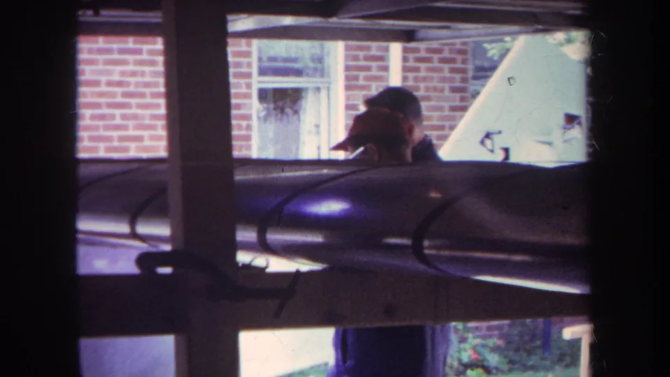 1969 Yucaipa home-movie frame showing a man in a cap outside a brick house, partly blocked by window mullions and an aircraft wing or fuselage.