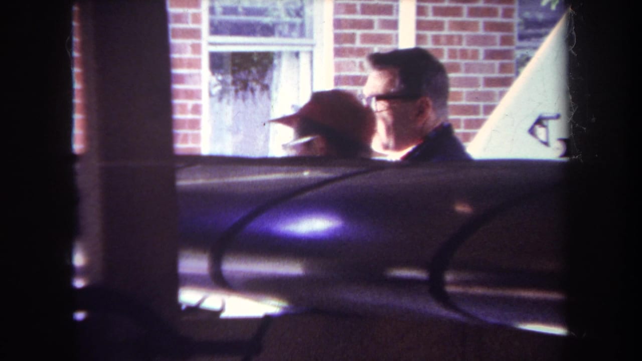 Man in red hat and man with glasses stand outside a brick house behind aircraft parts.