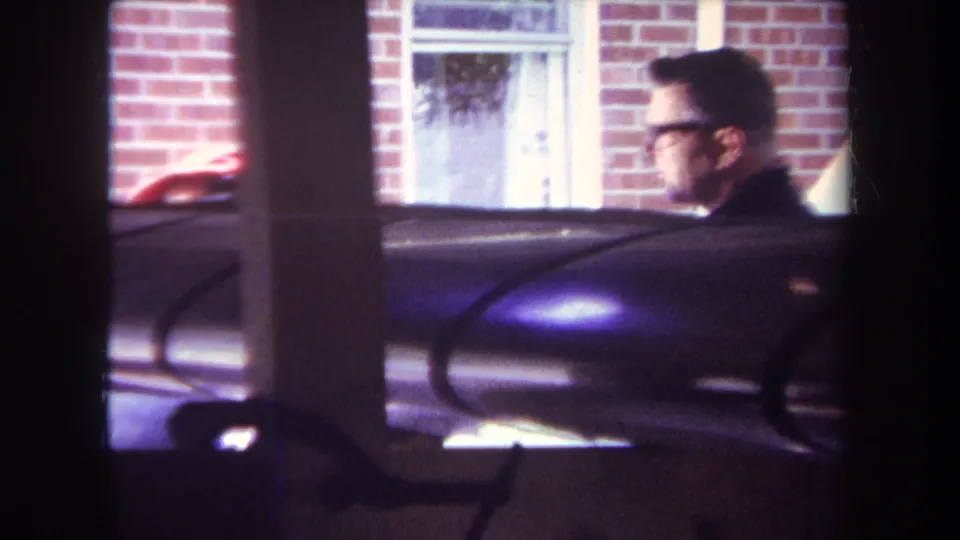 1969 Yucaipa home-movie frame showing a man with glasses moving past a brick house, with dark window mullions and aircraft parts in the foreground.