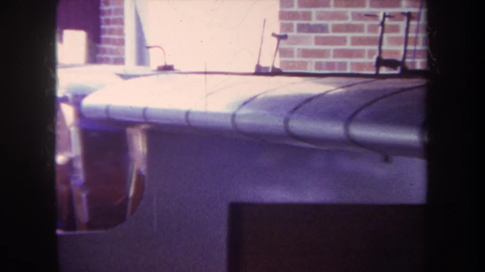 1969 Yucaipa frame showing the side of a silver model airplane with canopy detail beside a brick wall and bright background opening.