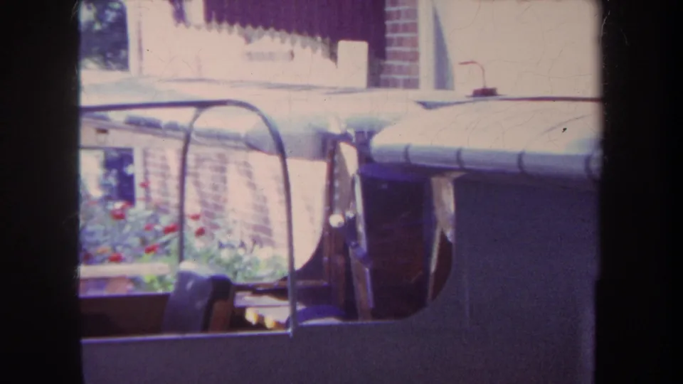 1969 Yucaipa home-movie frame showing the model airplane canopy with garden flowers, foliage, and brick wall in the background.