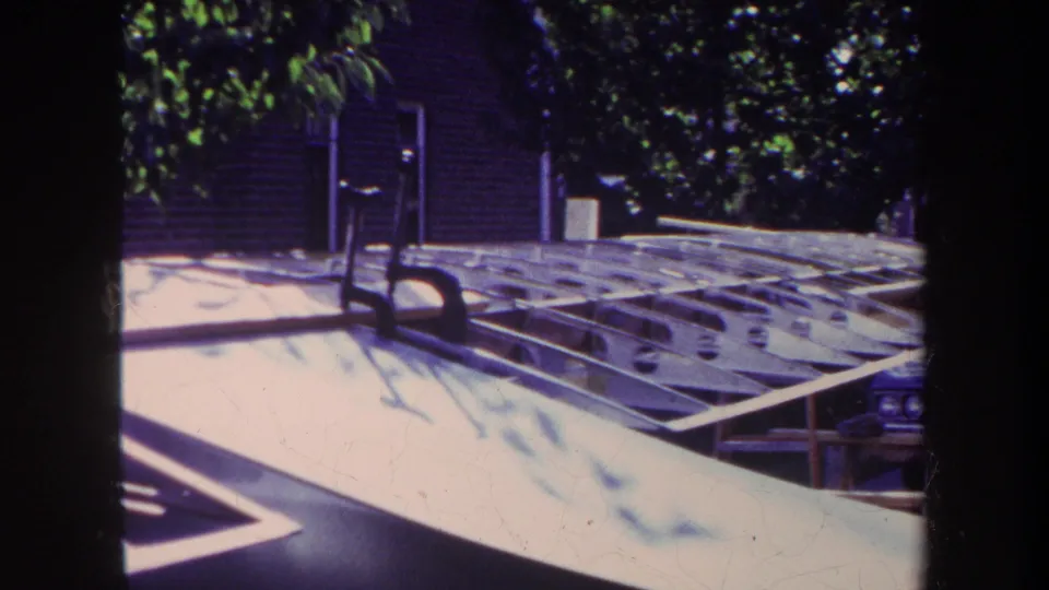 A 1969 home-movie view of the homemade aircraft under construction, with wooden framing, a pale surface, trees, and a residential building behind it.