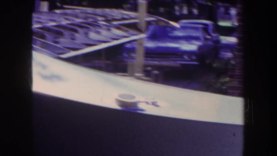 A closer 1969 frame shows the pale aircraft body or wing edge, cockpit opening, exposed ribs, support posts, and a dark car behind the build.