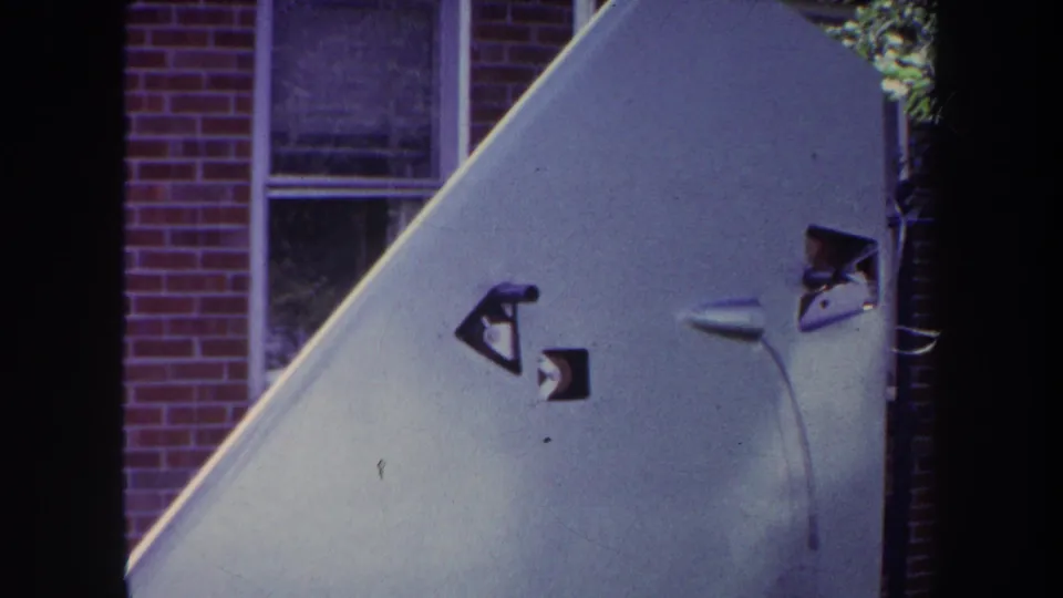 1969 Yucaipa frame of a pale aircraft tail surface with dark decals, a small mounted part, and a brick house background.