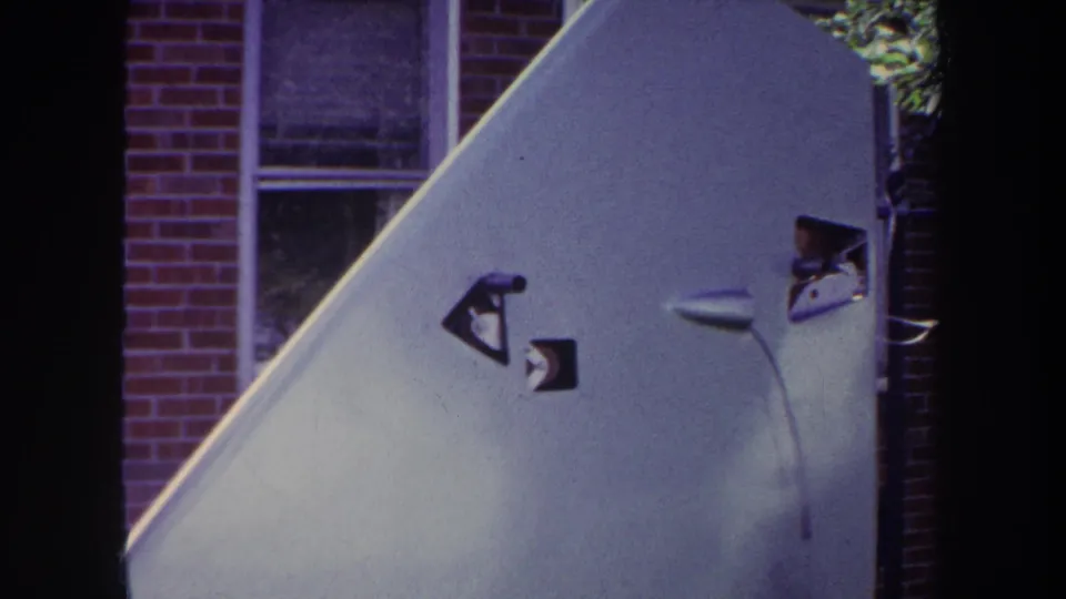 1969 Yucaipa home-movie still showing the aircraft tail fin lower in frame with brick house windows in the background.