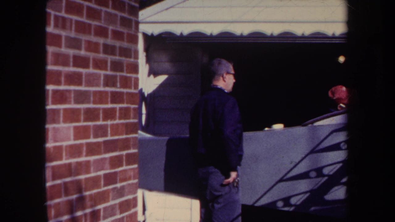In 1969 Volmer, Yucaipa home-movie footage, a man stands by a brick wall and garage while a person in a red cap is partly visible behind him.