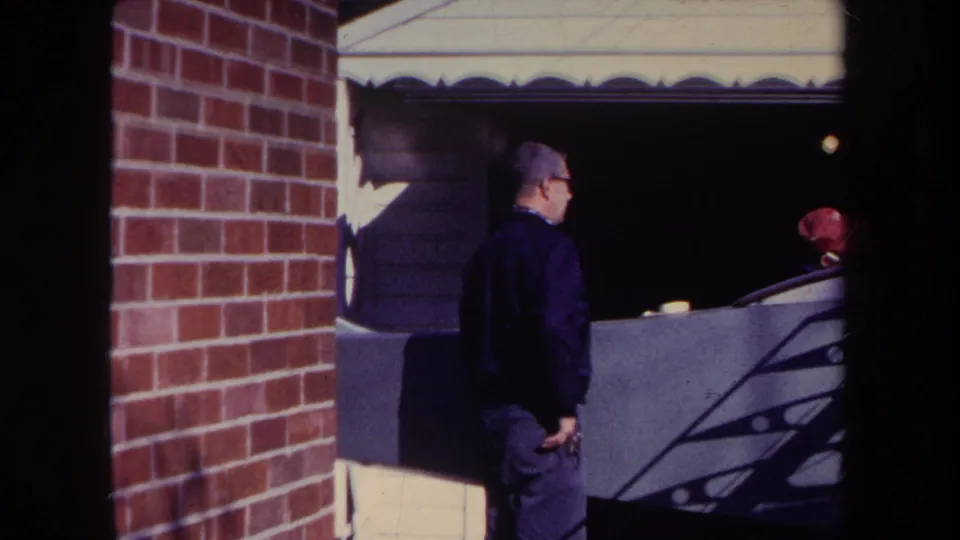 In 1969 Volmer, Yucaipa home-movie footage, a man stands by a brick wall and garage while a person in a red cap is partly visible behind him.