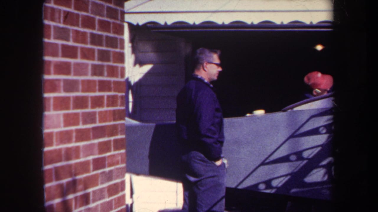Man walks or turns beside a brick wall and garage, with a person in a red cap behind him.