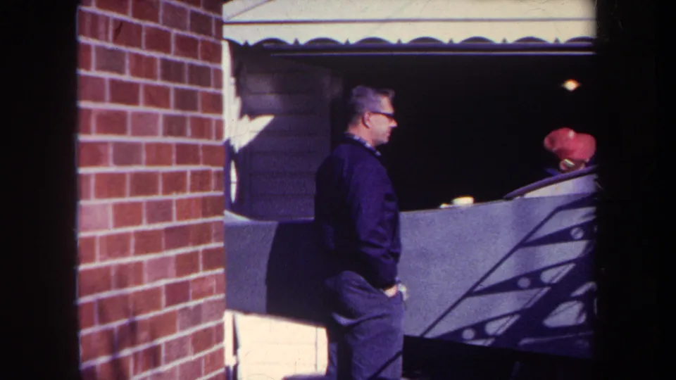 The man moves beside the garage exterior in the 1969 Yucaipa footage, with a second person in a red cap partly visible inside the shaded area.