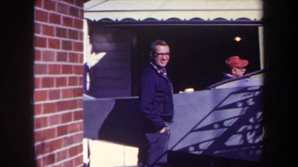 A man in glasses smiles toward the camera near a brick wall and garage in supplied 1969 Volmer, Yucaipa 8mm home-movie footage.