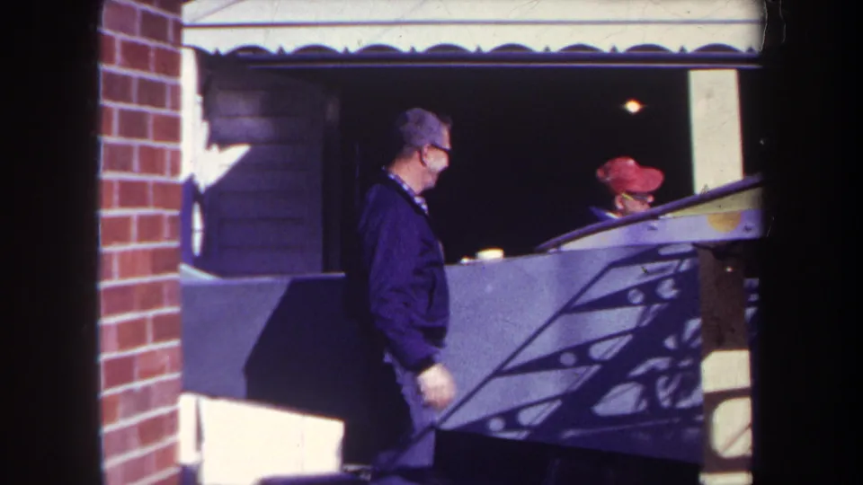Near the end of the 1969 Yucaipa clip, the man stands in profile by the garage as the red-capped person remains partly seen in the shaded opening.