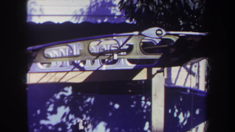 1969 home-movie still showing a close dockside view of a dark hull, pale frame, and diagonal rods on a small boat.