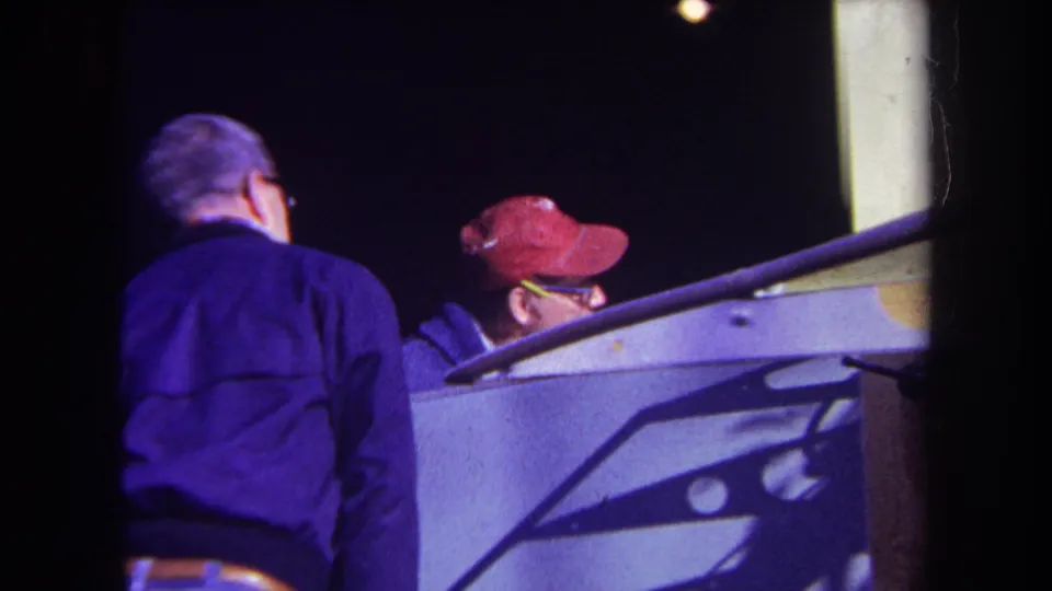 1969 Volmer Yucaipa frame showing two adult observers beside the unidentified mechanical structure, including one man in a red cap and glasses.