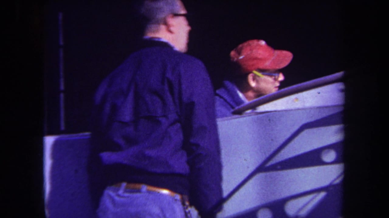 1969 Yucaipa home-movie frame showing two adult men, one in a red cap, beside a pale console-like piece of equipment at night.