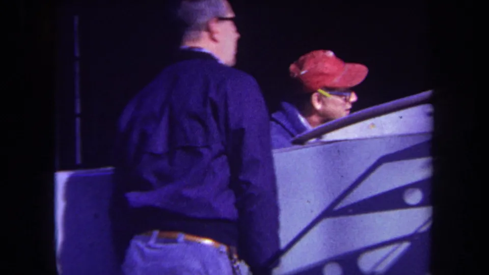 1969 Yucaipa home-movie frame showing two adult men, one in a red cap, beside a pale console-like piece of equipment at night.