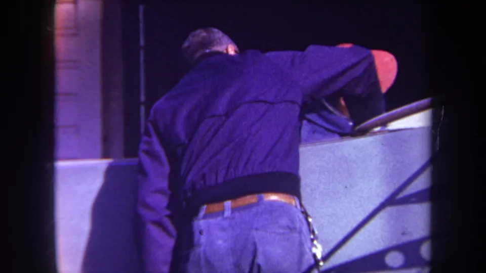 1969 Yucaipa frame with a man leaning into or over a pale console-like structure as another man in a red cap remains nearby.