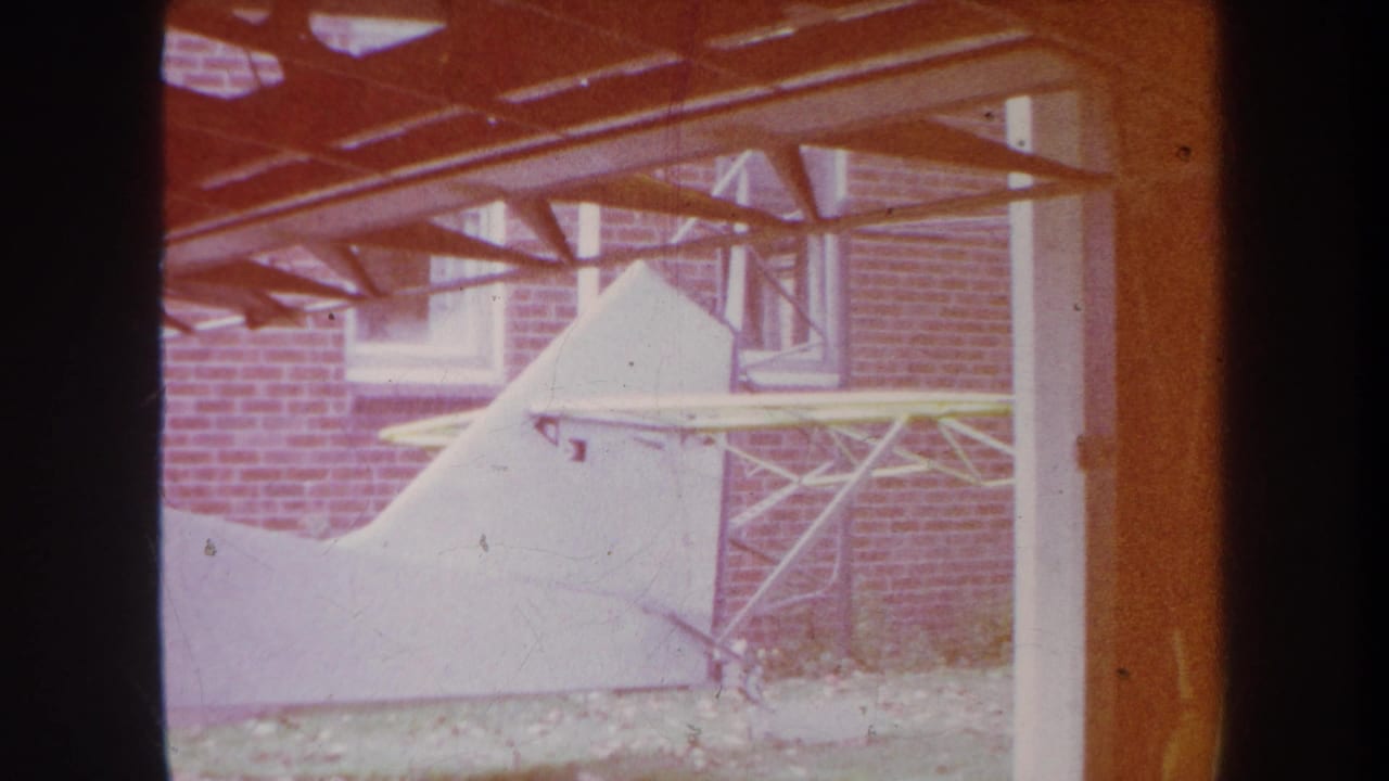 1969 8mm frame from Yucaipa showing a white aircraft tail or tail section near a brick building and overhead wooden beams.