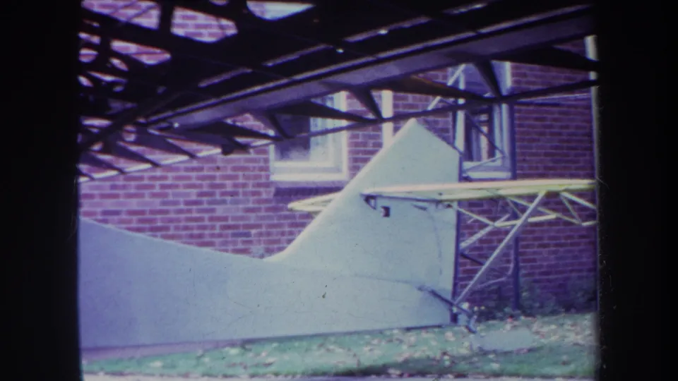 Closer 1969 home-movie view of a white aircraft tail or fuselage section under a timber structure near a brick building.